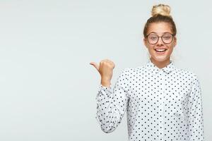 Portrait of happy attractive young woman with bun wears polka dot shirt and spectacles smiling and points to the side isolated over white background photo