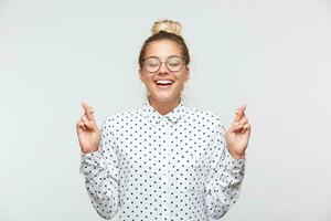 Closeup of cheerful young woman with bun and closed eyes wears polka dot shirt and glasses feels inspired, keeps fingers crossed and making a wish isolated over white background photo