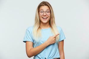 Closeup of cheerful surprised young woman with blonde hair wears spectacles and blue t shirt feels happy and points to the side with finger isolated over white background photo