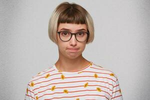 Portrait of shy sad cute young woman wears striped t shirt and glasses feels upset and pressing her lips isolated over white background photo