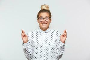 Portrait of happy excited young woman with bun wears polka dot shirt and spectacles keeps fingers crossed, biting lip and making a wish isolated over white background photo