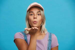 retrato de coqueta atractivo rubia joven mujer usa rosado gorra y Violeta t camisa mira juguetón y enviando aire Beso aislado terminado azul antecedentes foto