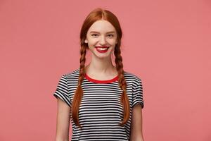 bonito bonito Pelirrojo niña con rojo labios, dos trenzas, encantador sonrisa, muestra blanco sano dientes, vestido en despojado camiseta, aislado en un rosado antecedentes foto