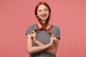 Indoor photo of pretty red-haired girl with red lips playing with two braids in hands dressed in stripped t-shirt, dreams imagines happy moments with closed eyes isolated on a pink background