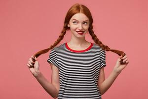 bonito bonito Pelirrojo niña con rojo labios participación dos trenzas en manos vestido en despojado camiseta, flirteador en broma mirando a el izquierda Superior esquina Graficado aventuras aislado en un rosado antecedentes foto