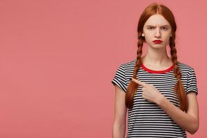 Serious displeased angry girl with two red haired braids red lip , dressed in stripped t-shirt, pointing fore finger to the upper left corner at blank copy space isolated on pink background photo