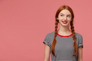 Chrarming pretty beautiful girl with red-haired braids red lips, dressed in stripped t-shirt, smiling dreamingly thoughtfully looks to the upper left corner stands next to the blank copy space photo