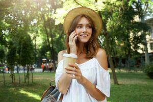 Outdoor portrait of happy cute young woman wears stylish summer hat, backpack and white dress, talking on cell phone and drinking takeaway coffee in park in the city photo