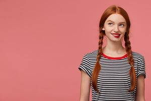 juguetón encantador niña con dos rojo peludo trenzas mordiendo rojo labio dentro tentación, vestido en despojado camiseta, soñando pensativamente mira a el Superior izquierda esquina soportes siguiente a el Copiar espacio foto