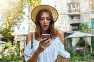 Outdoor portrait of amazed beautiful young woman wears stylish hat and white summer dress, feels surprised, using smartphone and drinking takeaway coffee in old city photo