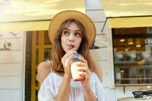 Outdoor portrait of thoughtful charming young woman wears stylish summer hat and white dress, feels relaxed, thinking and drinking milkshake in cafe in city photo