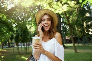 Outdoor portrait of amazed attractive young woman wears stylish summer hat and white dress, feels happy and surprised, walking in park with cup of takeaway coffee and talking on mobile phone photo
