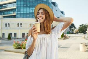 Outdoor portrait of cheerful charming young woman wears stylish summer hat and white dress, feels relaxed, standing and drinking takeaway coffee on the street in city photo