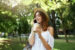 Outdoor portrait of happy cute young woman wears stylish summer hat and white dress, feels relaxed, smiling, talking on mobile phone and drinking takeaway coffee in park photo