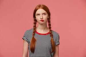 pensativo niña con dos rojo peludo trenzas mordiendo rojo labio dudas acerca de algo, vestido en despojado camiseta, mira a el Superior Derecha esquina aislado en rosado antecedentes foto