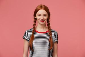 Attractive beautiful heartwarming pretty red-haired girl with red lips, two braids, chrarming smile, shows white healthy teeth, dressed in stripped t-shirt, isolated on a pink background photo