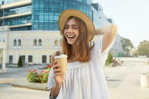 Outdoor portrait of cheerful beautiful young woman wears stylish summer hat and white dress, feels relaxed , drinks takeaway coffee and laughing on the street in city photo