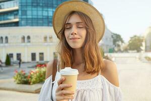 Outdoor portrait of happy beautiful young woman with eyes closed wears stylish hat, feels relaxed and drinking takeaway coffee in the city in summer photo