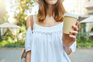 Cropped shot of beautiful young woman wears white summer dress holding cup of takeaway coffee and walking outdoors in old city photo