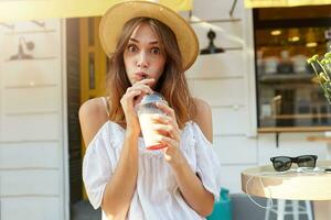 Outdoor portrait of amazed shocked young woman wears stylish hat and white dress , feels stunned and drinking milkshake in the city in summer photo