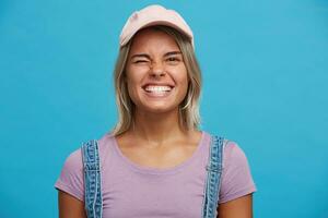 de cerca de alegre hermosa rubia joven mujer usa rosado gorra y Violeta t camisa mira feliz, sonriente y parpadeo aislado terminado azul antecedentes foto