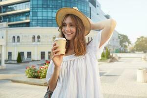 Outdoor portrait of smiling attractive young woman wears stylish summer hat and white dress, feels happy, walking in the city and drinks takeaway coffee photo