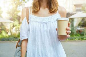 Cropped shot of attractive slim young woman wears white summer dress with cup of takeaway coffee walking outdoors in the park photo