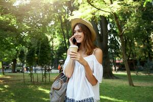 Outdoor portrait of happy beautiful young woman wears stylish hat, white blouse and striped backpack, feels relaxed, talking on mobile phone and drinking takeaway coffee in park in summer photo