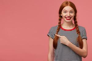 Close up of happy inspired red haired teen girl shows with index finger on the empty space on left side,is delighted with the product, advises to pay attention,place for your advertisement photo