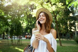 Outdoor portrait of cheerful attractive young woman with eyes closed holding cup of takeaway coffee, wears stylish hat, talking on cell phone and laughing in park in summer photo