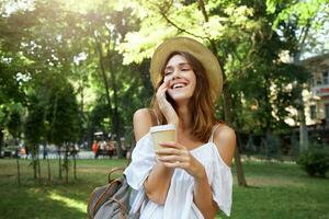 Outdoor portrait of smiling charming young woman wears stylish hat, backpack and white summer dress, holding cup of takeaway coffee, feels relaxed, talking on mobile phone in park in the city photo