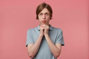 Young man with praying begging look looks upwards, arms up and fingers clenches fist while standing against pink background. Hoping, wishing, praying, expecting miracle photo