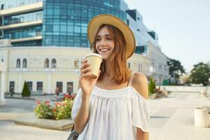 Outdoor portrait of happy cute young woman wears stylish summer hat and white dress, feels relaxed, smiling and drinking takeaway coffee on the street in city photo