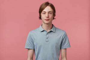 Young man did not slept for a long time, tired, zombie eyes, slightly-open eyes, looks calm, express no feelings, dressed in polo T-shirt, isolated over pink background photo