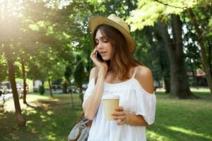 Outdoor portrait of beautiful young woman wears stylish hat, backpack and white summer dress, feels relaxed, talking on cell phone and drinking takeaway coffee in park photo