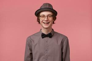 atractivo joven hombre inteligentemente vestido en camisa sombrero y negro corbata de moño usa lentes bien extensamente sonriente demostración ortodoncia soportes aislado en rosado antecedentes foto