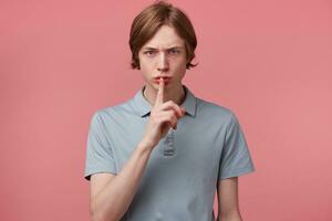 Young serious frowned guy keeps fore finger on lips, calls to keep a secret, do not tell anyone, keep quiet, do not make noise, demonstrates silence gesture, isolated over pink background photo