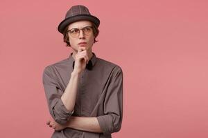 Young smartly dressed guy stands holding his hand on a chin, looking right upper corner thoughtfully, serious, thinking over a problem, ponders about some literature, over pink background photo