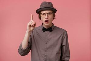 Serious young smartly dressed guy holding hand up pointing index finger upwards, looking camera through glasses moralizing, comment on issues of right and wrong, makes moral lecture, pink background photo