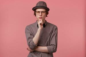 Serious young smartly dressed guy stands holding his hand on a chin, looking camera through glasses thoughtfully, worried, thinking over a problem, ponders about some literature, over pink background photo