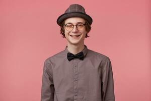 Young man in shirt hat and black bowtie wears glasses nice widely smiling showing orthodontic brackets isolated on pink background photo