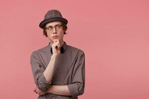 Doubtful young smartly dressed guy stands holding his hand on a chin, looking right upper corner thoughtfully, worried, thinking over a problem, scared of something, over pink background photo