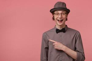 Elegant young guy opened his mouth in excitement, is overwhelmed with positive emotions happiness joy isolated on pink, pointing index finger to the left on copy space photo