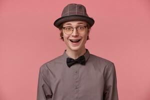 Excited surprised attractive young guy smartly dressed in shirt hat and black bowtie wears glasses, opened mouth because of amazement, has brackets, isolated on pink background photo