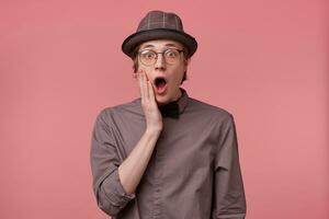Young guy seriously shocked very amazed with opened mouth touching face smartly dressed in shirt hat and black bowtie wears glasses feels surprised isolated on pink background photo