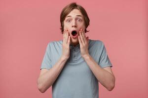 Close up of young man with blown away, amazed expression and dropped jaw isolated on pink background. Guy is shocked became a witnessed of the accident., hands touching face photo