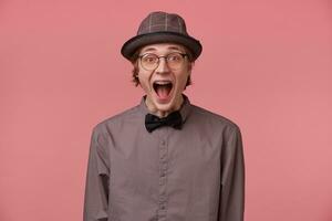 Guy opened his mouth in surprise, is overwhelmed with positive emotions happiness joy doesn't believe in his success, luck, dressed in shirt hat and black bowtie glasses has brackets isolated on pink photo