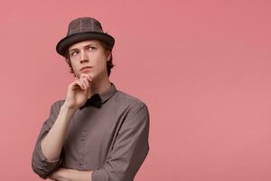 Young elegantly dressed guy stands holding his hand on a chin, looking right upper corner thoughtfully, serious, thinking about art, ponders about some literature, over pink background photo