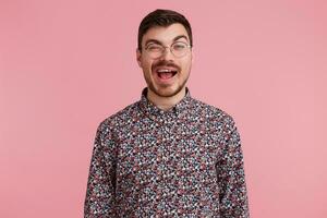 Close up photo of sweet heartbreaking darkhaired attractive man in glasses, flirting with someone, winks showing approval predisposed interes, in coloful shirt, isolated over pinkstudio background