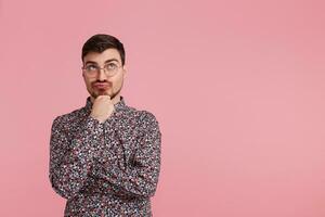 Young man in colorful shirt looking upwards, copy space on the right side,having doubts and with confuse face expression while scratching his chin isolated on pink background photo